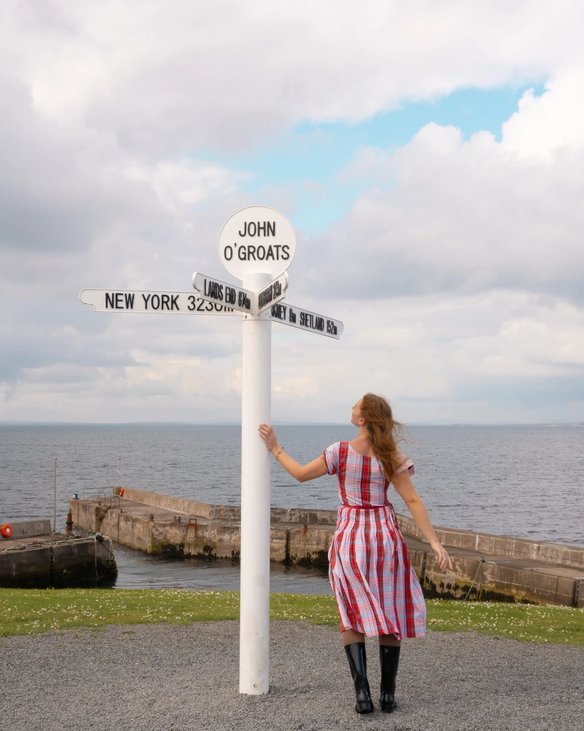 girl stands in a tartan dress at the signpost at the very top of scotland